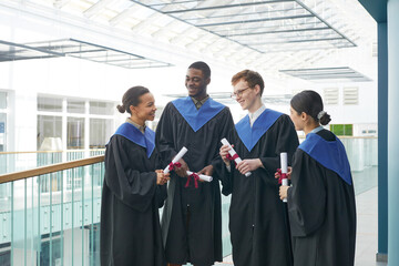 Fototapeta premium Diverse group of young people wearing graduation gowns chatting happily indoors in modern university interior