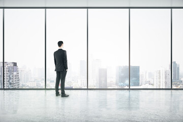Vision concept with businessman looking through big window on city skyscrapers in empty office hall with glossy floor.
