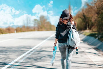 The concept of hitchhiking trips and local travel. Hipster woman in a cap, with backpack walking down the road holding a paper map in her hand. Blurred road on the background. Rear view