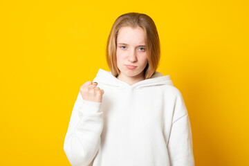 Do not mess with my anger. Annoyed girl clenches fist and looks irritated, dissatisfied with something, threatens to revenge, wears white hoodie, isolated on yellow background.