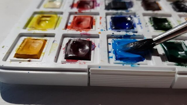 A set of used watercolor paints in a white case. Brush picks up blue paint.