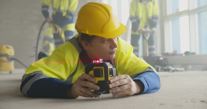 Builder in uniform lying on floor using laser level at construction site