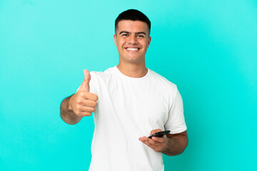 Young handsome man over isolated blue background using mobile phone while doing thumbs up