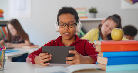 Cheerful african-american boy in headphones holding tablet while sitting at desk in school classroom
