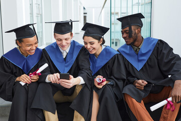 Diverse group of smiling young people wearing graduation gowns and using smartphone while sitting on steps outdoors