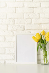 yellow tulips in a glass vase and blank photo frame on a white brick wall background