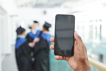 Close up of African-American man holding smartphone with blank screen to camera with group of college graduates in background, copy space