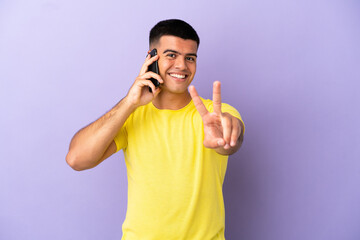 Young handsome man using mobile phone over isolated purple background smiling and showing victory sign
