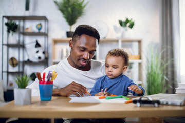 Obraz premium Handsome afro american man holding his cute son on knees and drawing with colorful pencils. Young father sitting at table with modern laptop and entertaining child.