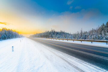 Obraz premium Sunrise on a clear winter morning, an empty country road to snowfall passing through a coniferous forest. View from the side of the road