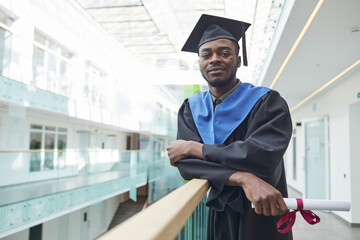 Waist up portrait of African-American young man wearing graduation gown and hat looking at camera...