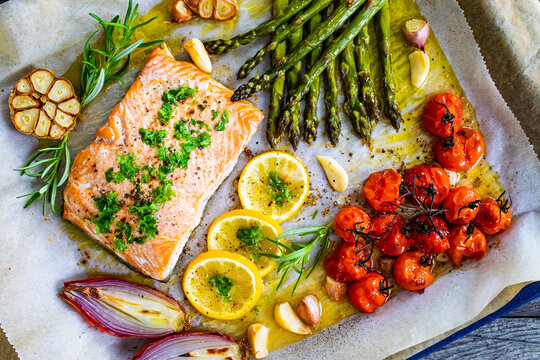 Sheet pan dinner - roasted salmon steak with asparagus, lemon ,rosemary, tomatoes, onion and garlic on cooking pan on wooden table
