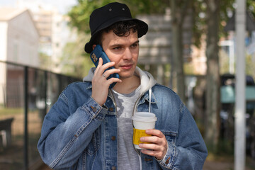 Fototapeta premium Young handsome man in a denim jacket drinking coffee from a paper cup and talking on the phone. Happy man on a phone call walking on a sunny day. 