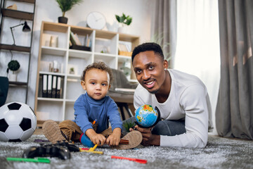 Joyful afro american man and little baby boy playing with toys on carpet, smiling and looking at camera. Leisure time for family at home.