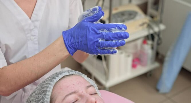 Cosmetologist hands in blue gloves touching face of caucasian teen girl patient making cleaning peeling procedure in beauty salon. Acne, skin treatment, problem skin. Skin care concept. Face wash