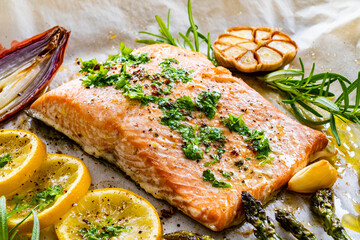 Sheet pan dinner - roasted salmon steak with asparagus, lemon ,rosemary, tomatoes, onion and garlic on cooking pan on wooden table

