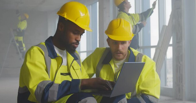 Portrait of two diverse builders reading plan of building on laptop