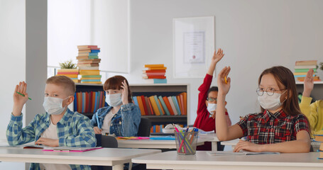Diverse children raising hands for answer wearing medical masks for protection from virus at lesson