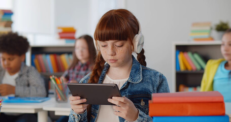 Portrait of cute redhead preteen girl in headphones using digital tablet at classroom
