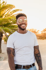 Portrait of a handsome black and young american man wearing sunglasses and smiling during sunset at the beach