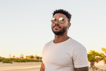 Portrait of a handsome black and young american man wearing sunglasses and smiling during sunset at the beach