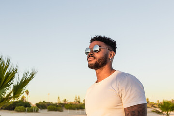 Portrait of a handsome black and young american man wearing sunglasses and smiling during sunset at the beach