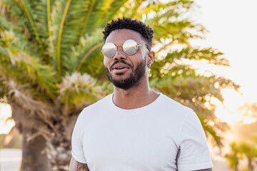 Portrait of a handsome black and young american man wearing sunglasses during sunset at the beach with palms in the background
