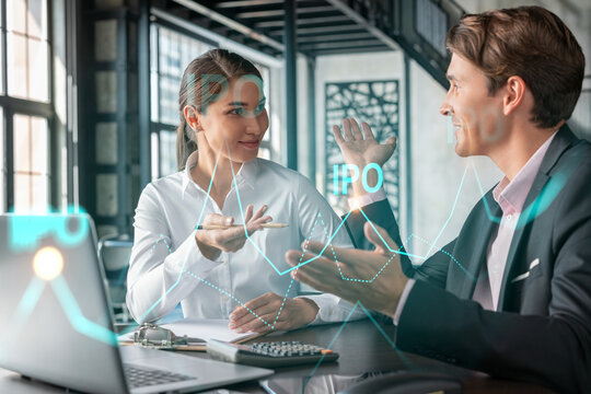 Businessman and businesswoman in formal wear working together to optimize IPO strategy at corporate fund. Financial chart hologram over office background with panoramic windows