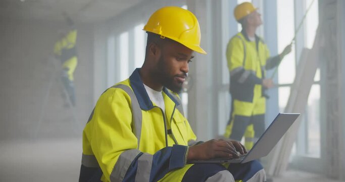 Portrait of handsome afro-american builder in helmet sitting with laptop at construction site