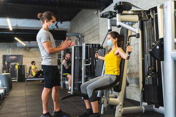 Fototapeta na wymiar Personal trainer helping young female athlete in the gym while both of them wearing protective face masks
