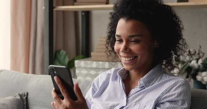 Close up attractive young 35s African woman sit on sofa with cell phone. New freeware mobile application usage, e-date services for singles, social media everyday usage, modern tech overuse concept