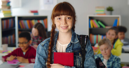 Portrait of pretty redhead schoolgirl holding book and smiling at camera standing in classroom