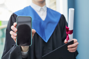 Close up of unrecognizable college graduate holding smartphone with blank screen to camera during ceremony, copy space