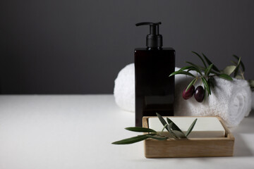 A black jar with a dispenser, a white towel, soap, a sprig of an olive tree with olives are on a gray background. Natural cosmetics theme. Copy space.