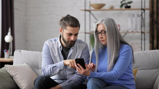Adult son teaching his senior mother to use smartphone mobile apps, sharing photos in social media, shopping in online store. Two generations family spending time together at home.