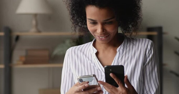 African smiling woman standing indoor holding in hands two cell phones, makes data transfer between new and used gadget. Synchronization of smartphones process, happy user of modern devices concept