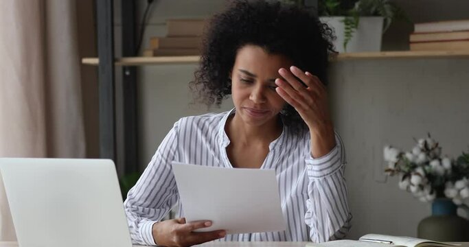African woman sit at workplace desk holding papers reading bad news in letter feels frustrated concerned due high taxes, loan debt, dismissal, staff cuts notice, debt eviction notification concept