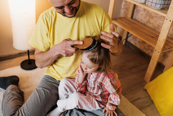 a caring father combs his little daughter's hair while sitting on the floor at home.