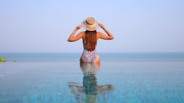 Back of a woman in swimwear sitting on infinity pool border, touching her sun hat and raising hands up with a stunning view of tropical sea horizon in Miami, Florida, static