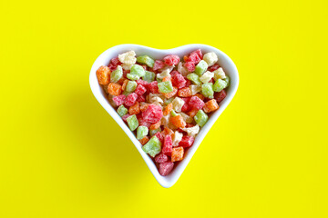 Candied fruits multicolored on a platter in the shape of a heart on a yellow background top view. 