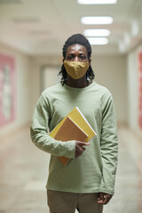 Vertical portrait of young African-American man wearing mask in school corridor and looking at camera