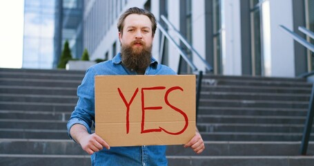 Portrait shot of Caucasian male activist with beard holding poster Yes at demonstration or protest. Single protest outdoor. Activism concept. Man showing table with word yes.