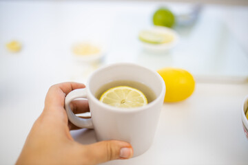 Female hand holding a cup of lemon tea above limes and lemons
