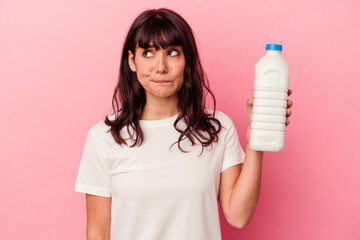Young caucasian woman holding a bottle of milk isolated on pink background confused, feels doubtful...