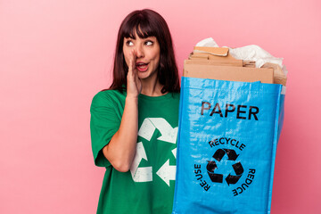 Young caucasian woman holding a recycled cardboard bag isolated on pink background is saying a secret hot braking news and looking aside