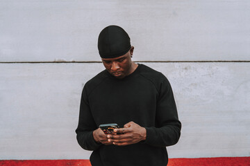 Chico negro vestido completamente de negro mirando smartphone frente a pared blanca