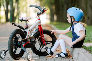 Cheerful smiling girl in helmet sitting near bicycle. Kid checking bike tires