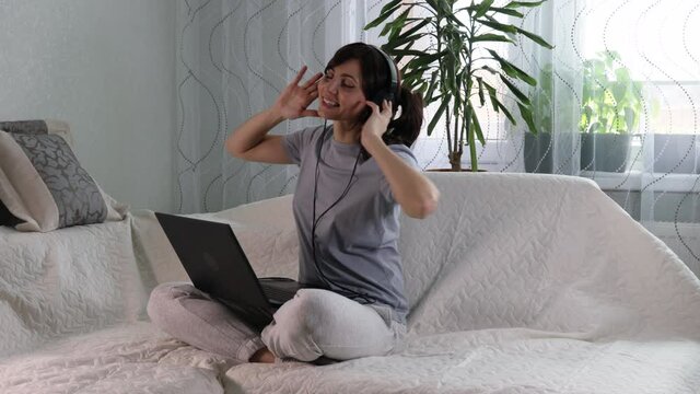 Attractive european woman with long dark hair in headphones in cozy interior listening music and dancing with hands sitting on comfy sofa at home near laptop computer. Relaxing time, Isolation, rest 