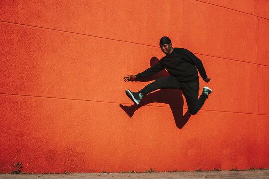Alegre varón español negro en ropa casual negra y do-rag saltando contra una pared roja