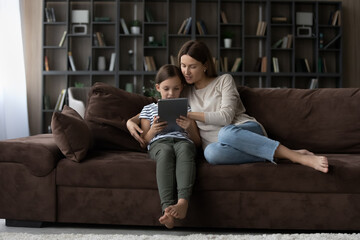 Loving young mother and teen daughter sit on sofa in living room study learn together online on tablet gadget. Caring mom and little teenage girl child use pad device at home. Technology concept.